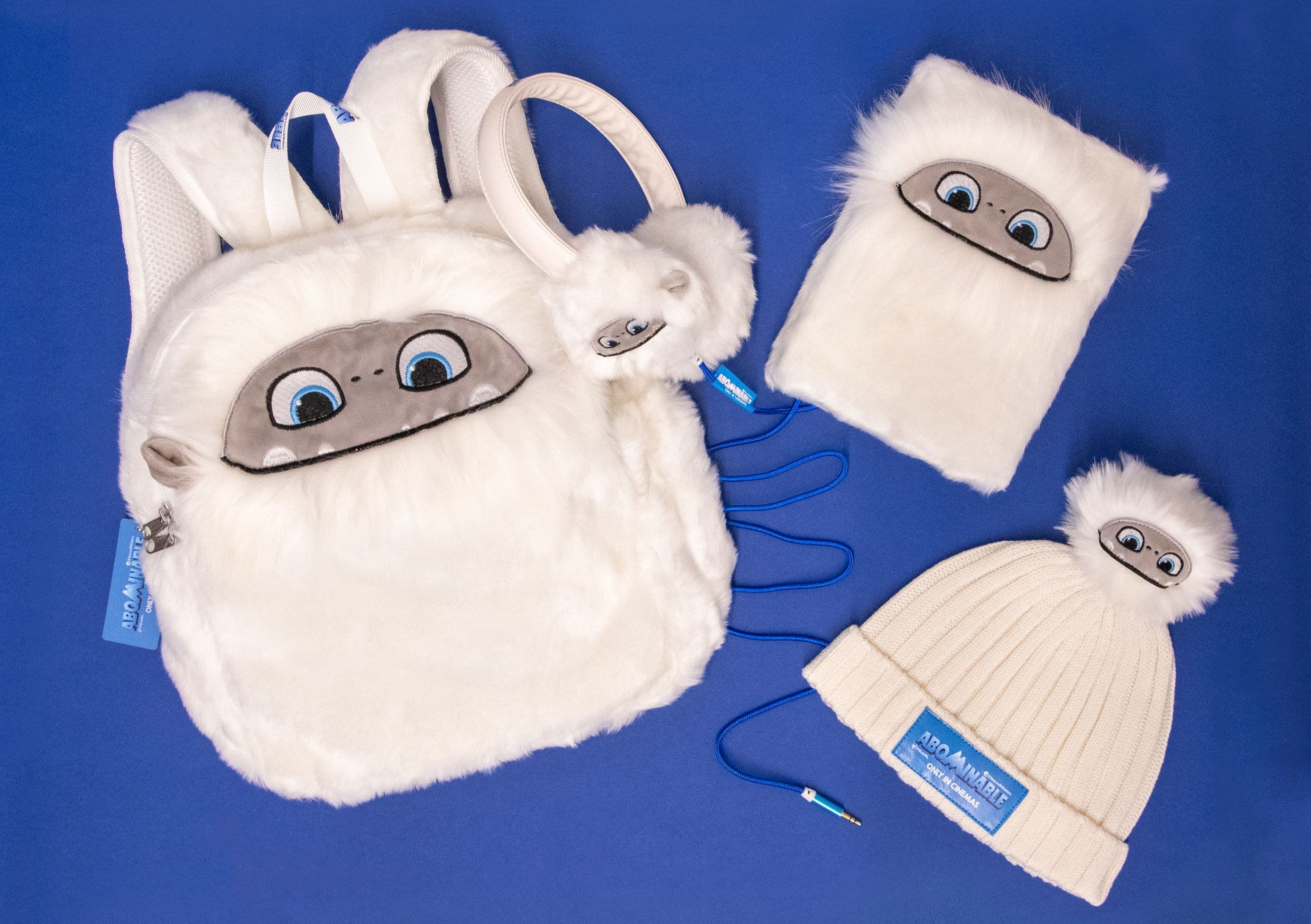 Abominable range on a blue background: a white plush yeti backpack, matching plush headphones, a fur-fronted notebook, a bobble hat and a character keyring